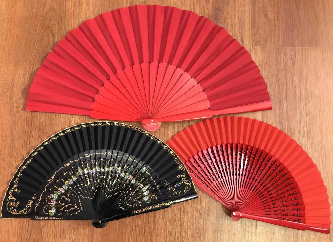 Tres abanicos: uno negro decorado y dos rojos, mostrando las partes de un abanico desplegado sobre madera.