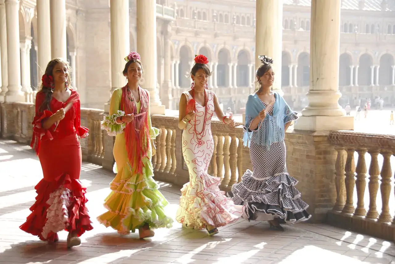 Cuatro mujeres con trajes de danza sevillana coloridos y volantes, pasean sonrientes bajo arcos.