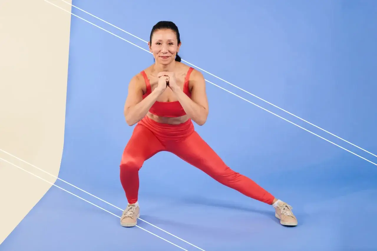 Mujer en ropa deportiva roja haciendo sentadilla lateral, estirando la parte interna del muslo.