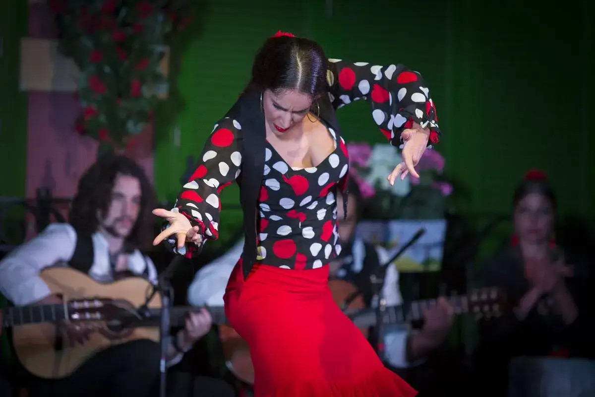 Bailarina de flamenco con falda roja y blusa de lunares, moviendo las manos al ritmo de las seguidillas. Guitarristas acompa&ntilde;an la actuaci&oacute;n.