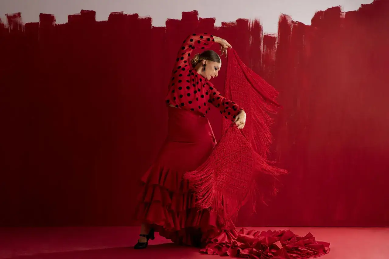 Una flamenca bailando con un vestido rojo de lunares negros y un mant&oacute;n de flecos, moviendo su cuerpo con pasi&oacute;n.