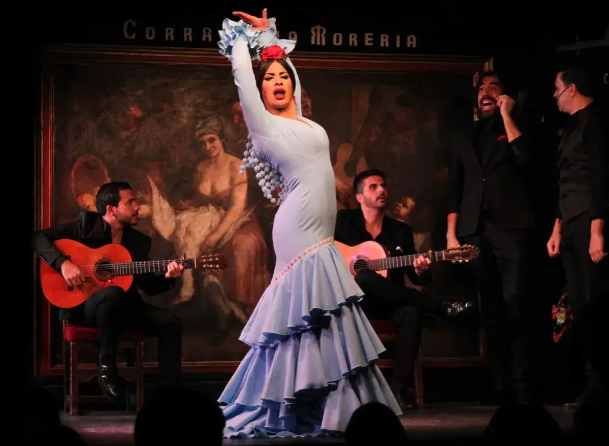 Una bailaora de flamenco, con un vestido azul voluminoso, expresa pura alegr&iacute;a en el escenario, acompa&ntilde;ada por guitarristas.