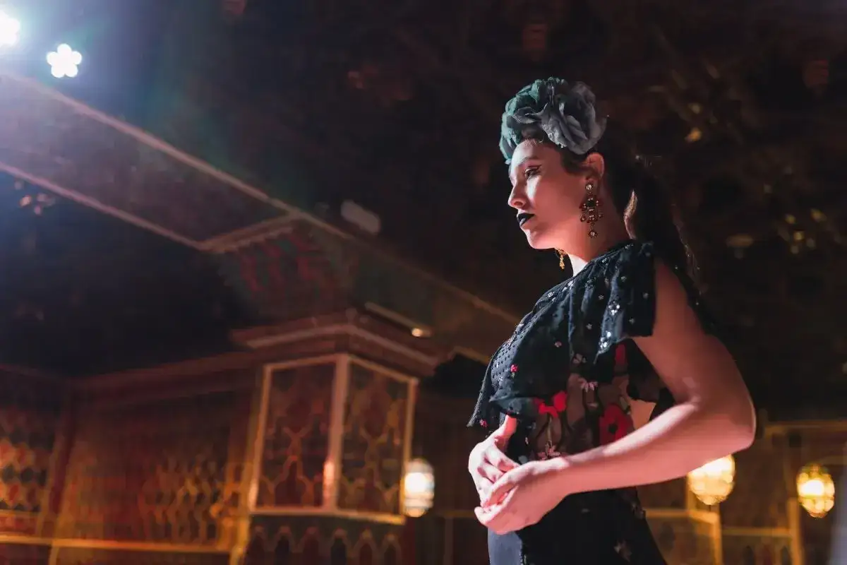 Una mujer con un gran adorno floral en la cabeza y maquillaje oscuro, vestida con un traje de volantes, parece una duende que es, posando en un escenario ornamentado.