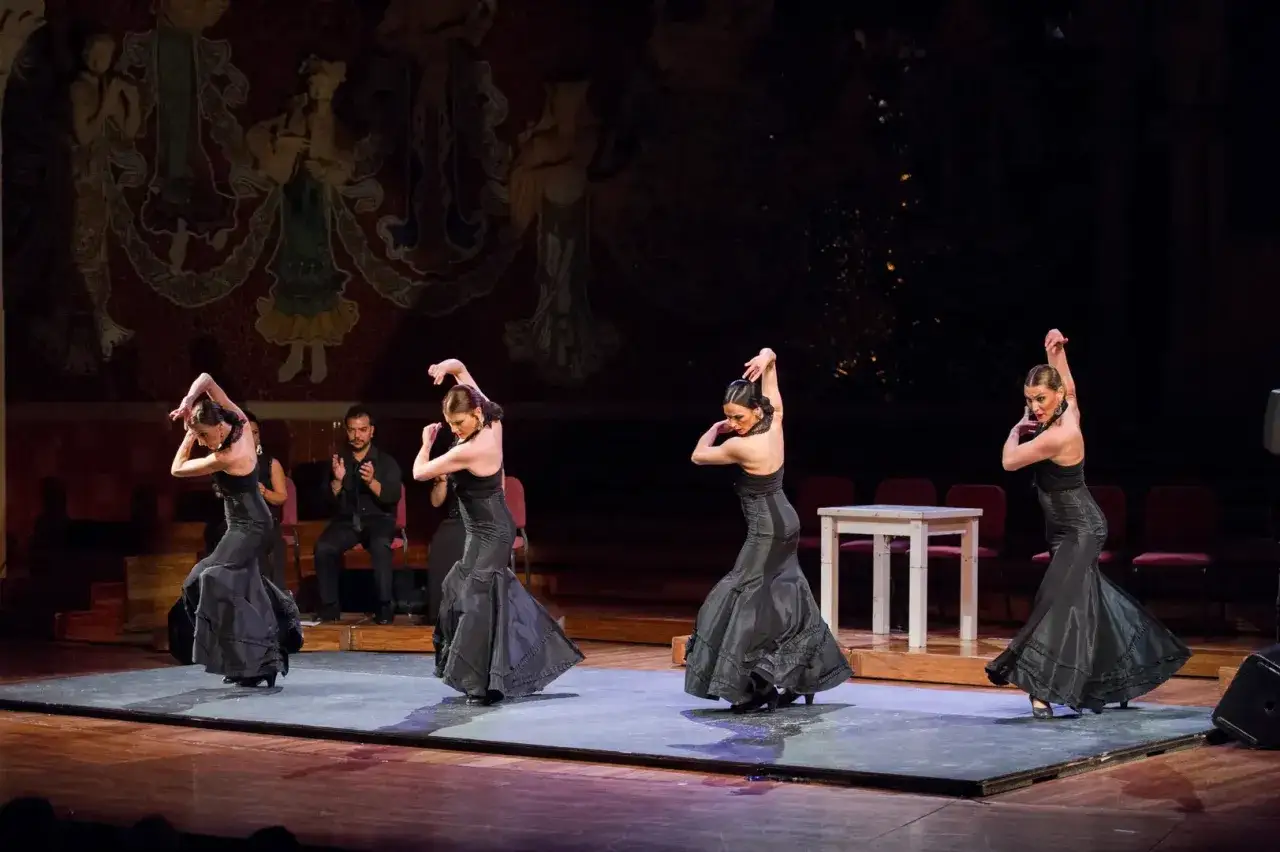 Cuatro bailarinas de flamenco, vestidas de negro, ejecutan un jaleo flamenco en el escenario. Un hombre aplaude al fondo.