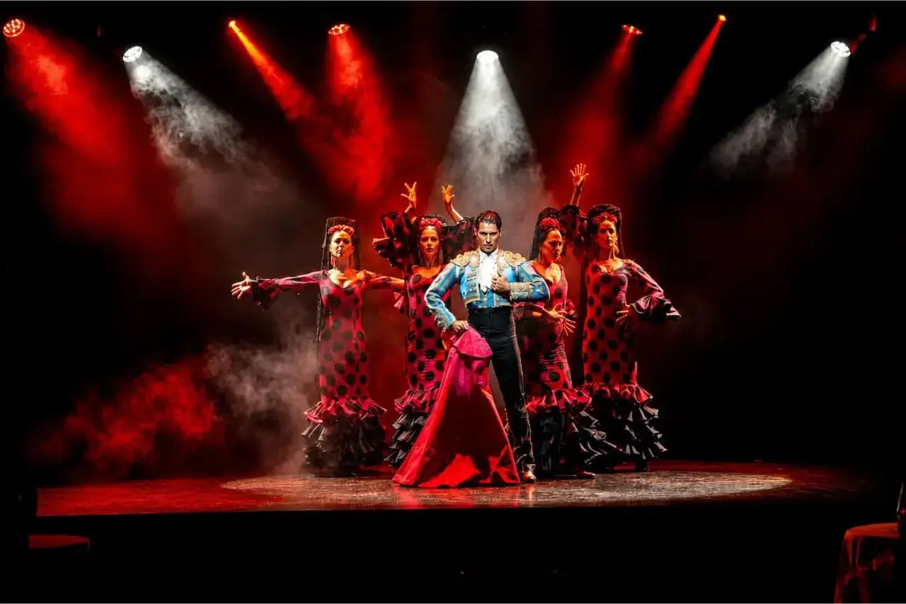 Un hombre y cinco mujeres en trajes de flamenco danse posan en un escenario con luces rojas y humo.