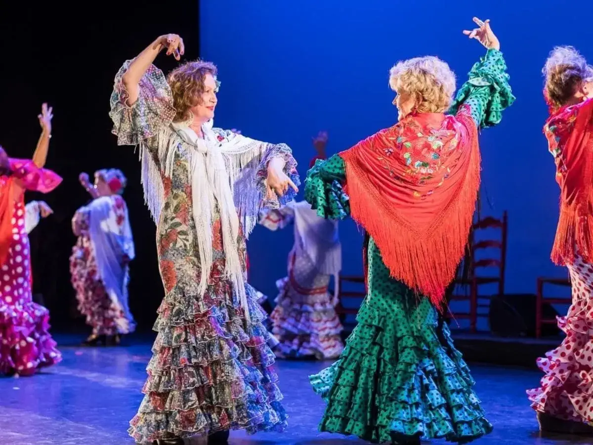 Dos mujeres bailan sevillanas y flamenco con trajes coloridos y volantes.