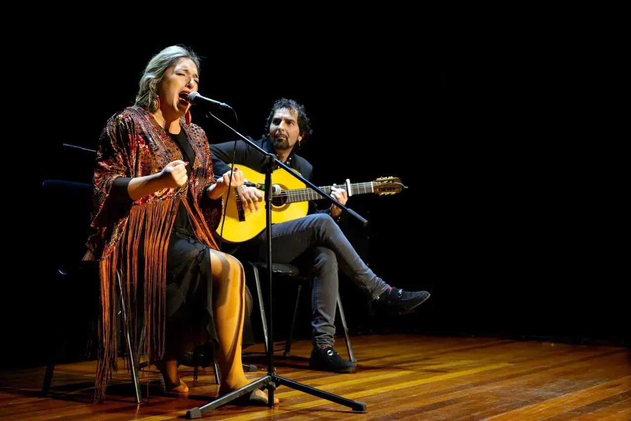 Una mujer canta apasionadamente frente a un micrófono, mientras un hombre la acompaña con la guitarra. El sonido flamenco llena el escenario.