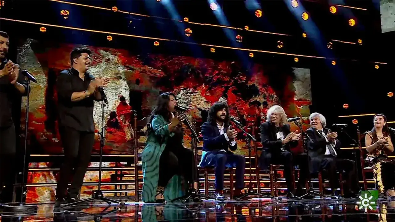 Un grupo de artistas, incluyendo un cantaor gitano, aplauden en un escenario con luces y un fondo abstracto.