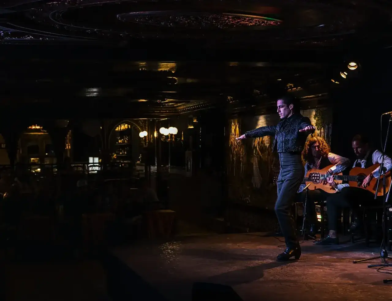 Un bailaor espa&ntilde;ol, con brazos extendidos, cautiva en el escenario. Dos guitarristas lo acompa&ntilde;an, creando una atm&oacute;sfera flamenca.