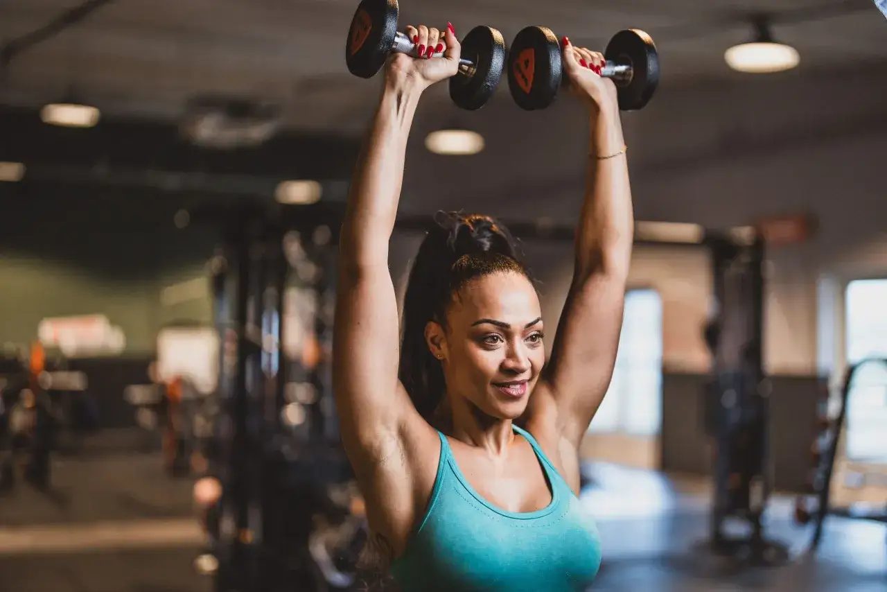 Mujer sonriente realiza ejercicios fitness con pesas en el gimnasio, levantando mancuernas sobre su cabeza.