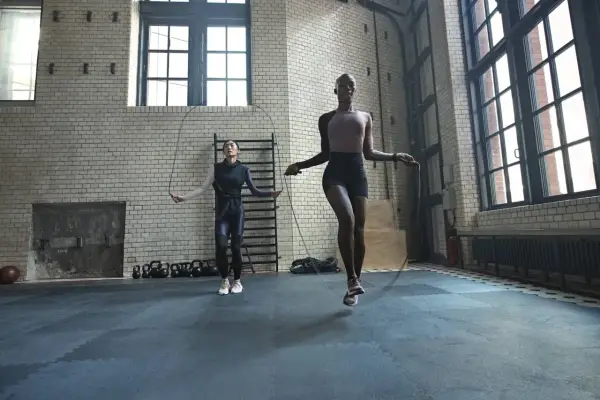 Dos mujeres realizan ejercicios de calentamiento general saltando la cuerda en un gimnasio industrial.