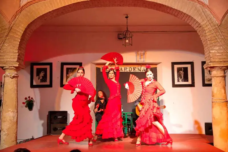 Tres bailarinas de flamenco, vestidas de rojo y con abanicos, ejecutan fandangos flamencos en un escenario.